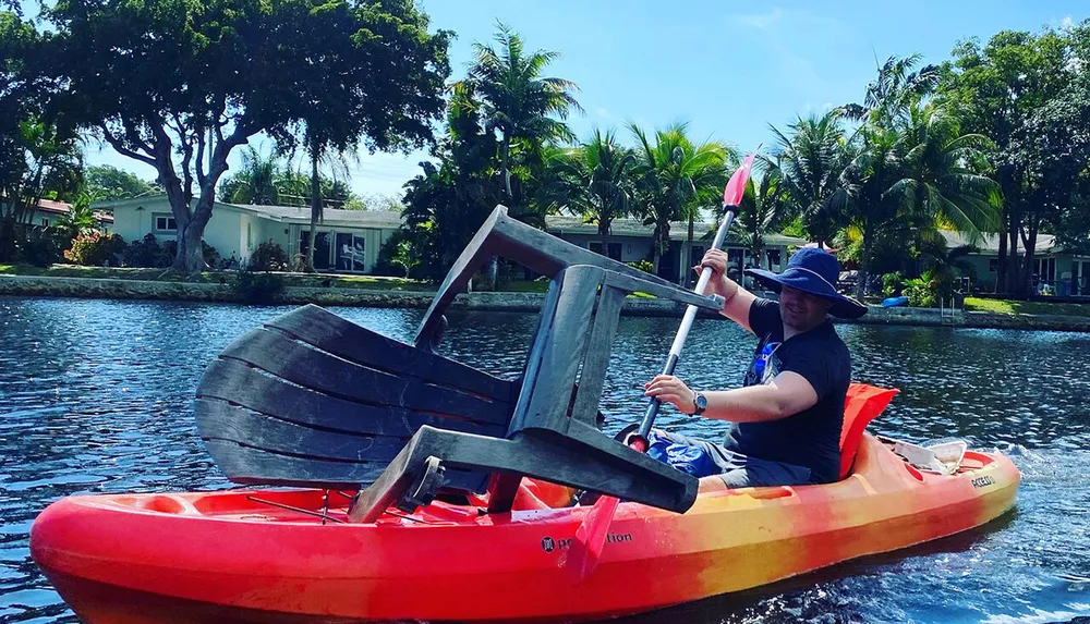 A person is kayaking with a large wooden chair strapped to the front of their kayak on a sunny day in a suburban waterway