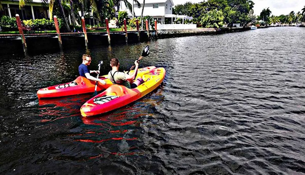 Two people are kayaking on a calm river with lush greenery and residential buildings on the sides