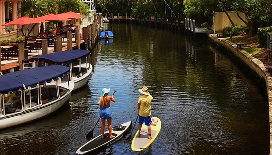 Two people are paddleboarding on a peaceful waterway flanked by boats and a dining area with red umbrellas.