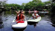 Two people are smiling and posing on stand-up paddleboards on a tranquil river surrounded by lush greenery.