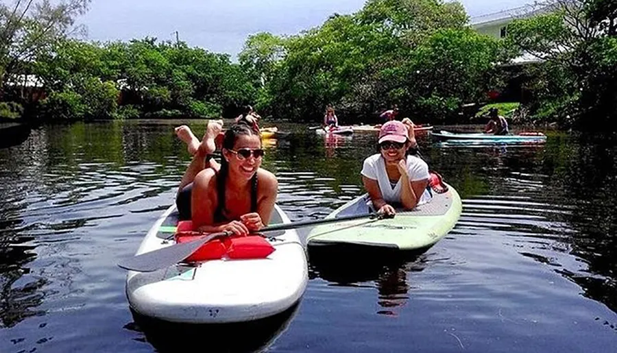 Two people are smiling and posing on stand-up paddleboards on a tranquil river surrounded by lush greenery.