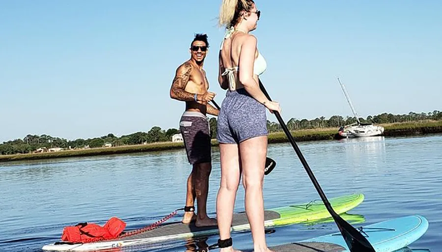 Two people are standing on paddleboards on a calm body of water, with a boat visible in the background.