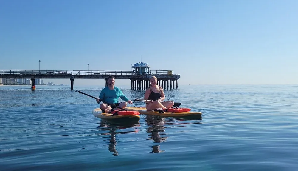 Two people are kayaking on calm waters with a pier in the background under a clear blue sky