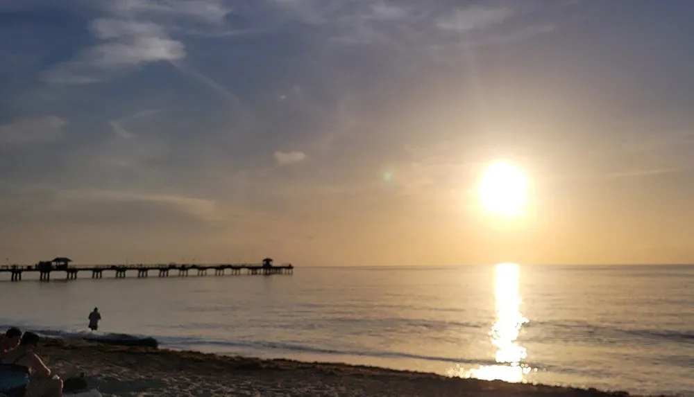 The image captures a serene sunset over the ocean with a pier in the distance and people enjoying the view from the beach