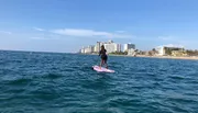 A person is paddleboarding on a calm sea with a view of a coastal city skyline in the background.