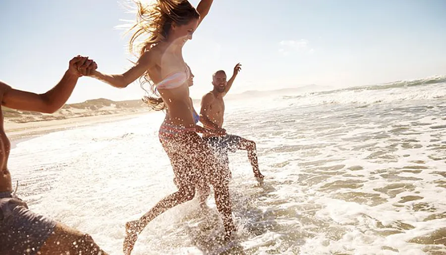 Three joyful people are holding hands and splashing through the surf on a sunny beach.