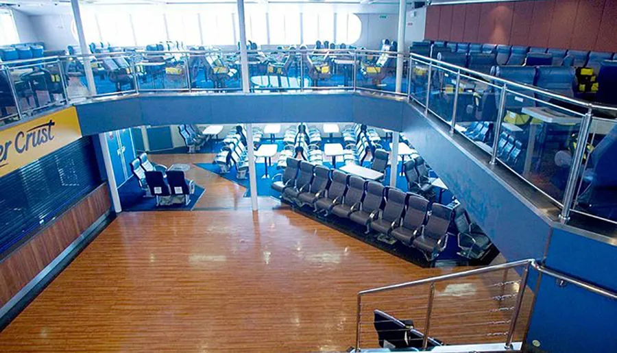 The image shows an empty ferry or ship interior with rows of seating and a closed snack bar, showcasing a modern and spacious design with two levels.