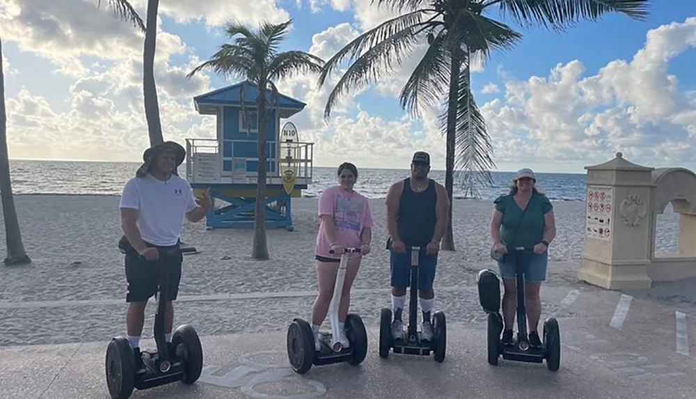 Four people are standing on Segways at a beach promenade with palm trees and a lifeguard tower in the background
