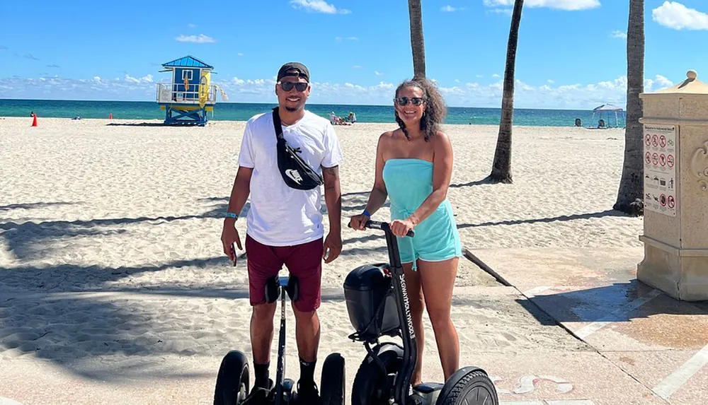 A man and a woman are smiling for the camera while standing next to a Segway on a sunny beach promenade with palm trees and a lifeguard hut in the background