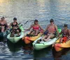 A group of smiling people are seated in colorful kayaks on calm water with two dogs also visible on the kayaks