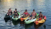 A group of smiling people are seated in colorful kayaks on calm water, with two dogs also visible on the kayaks.