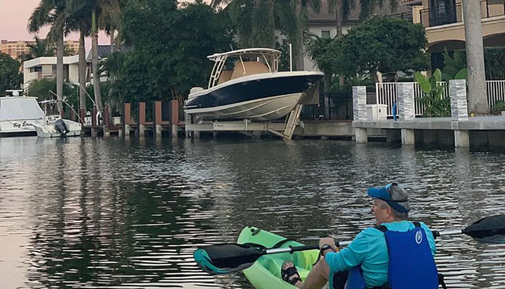 Two people are kayaking in water with a lifted boat docked in the background near residential buildings at dusk