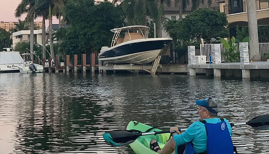 Two people are kayaking in water with a lifted boat docked in the background near residential buildings at dusk.