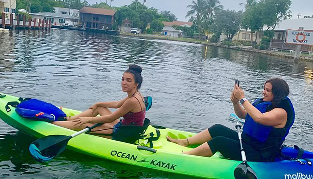 Two people are relaxing on a tandem kayak with one taking a photo while on calm waters