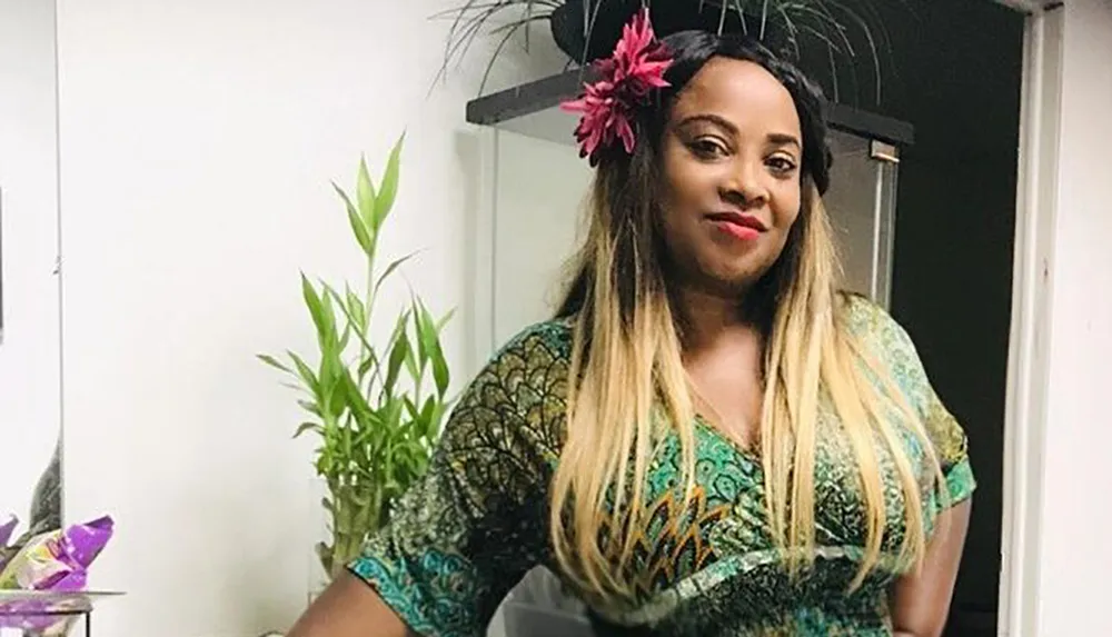 A person is standing indoors smiling at the camera wearing a patterned outfit with a pink flower in their hair near a green potted plant