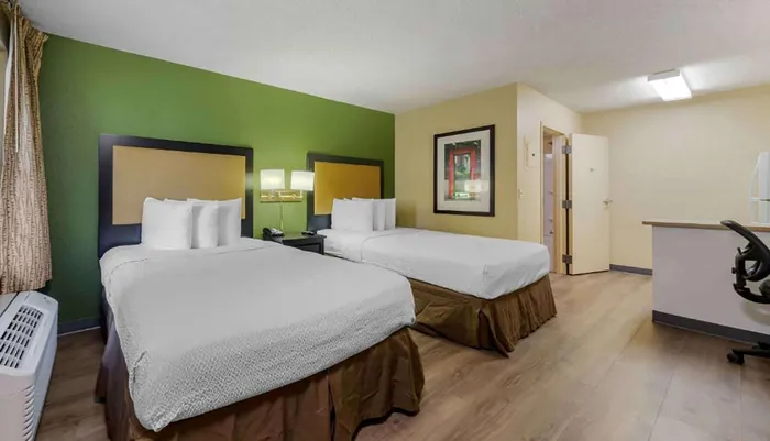 The image shows a neatly arranged hotel room with two beds a desk a framed picture on the wall and a green accent wall