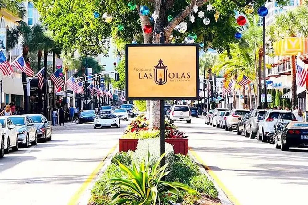 The image shows a vibrant street lined with parked cars and American flags with a sign reading Welcome to Las Olas in the foreground