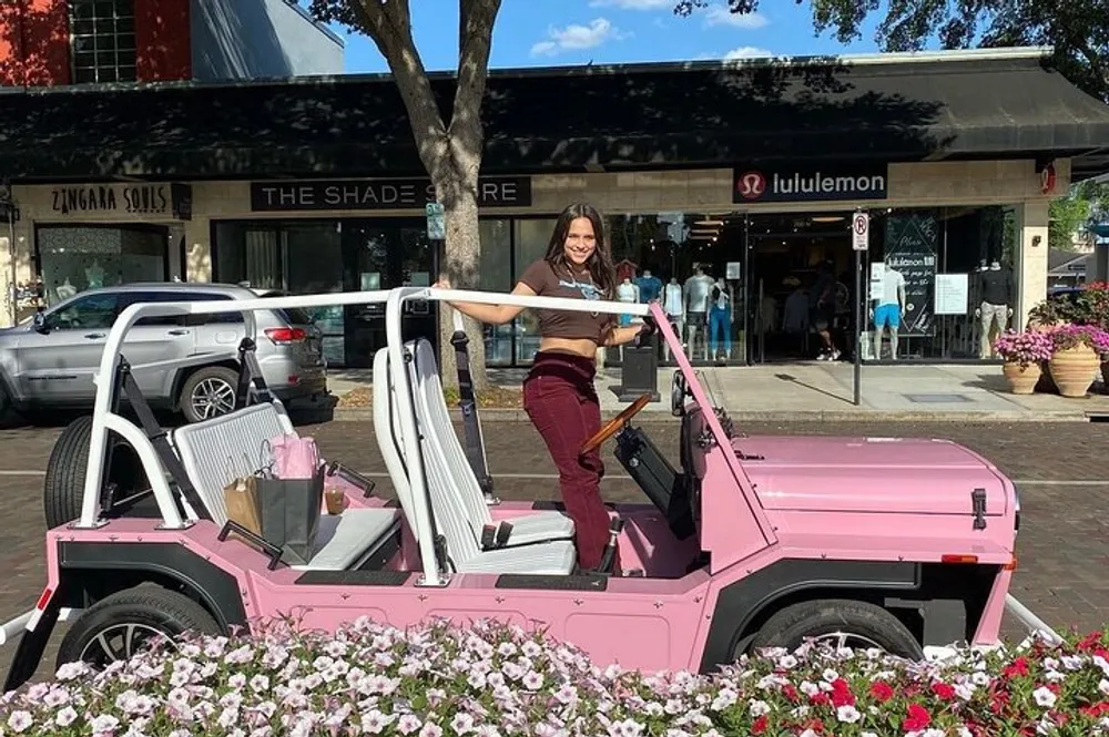 A smiling person is standing on the step of a pink doorless vehicle parked beside a flower bed on a sunny day in a shopping area