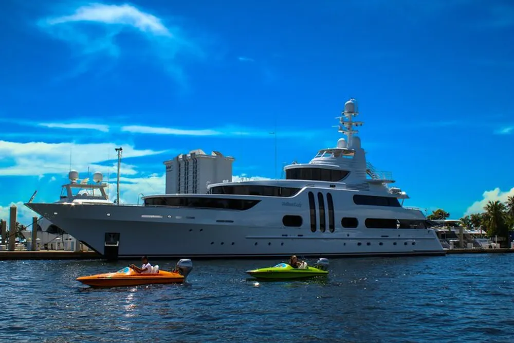A luxurious yacht is anchored on blue waters with two people on personal watercraft nearby under a partly cloudy sky