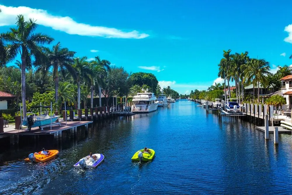A serene canal lined with palm trees and docks features colorful kayaks and luxurious boats under a bright blue sky