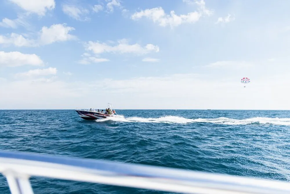 A speedboat moves across the blue sea with a parasailor in the sky viewed from another boats railing