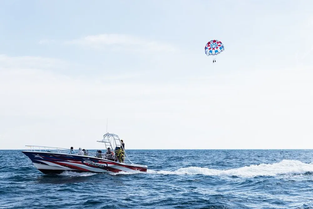 A speedboat is sailing on the ocean while a person parasails in the background under a colorful parachute