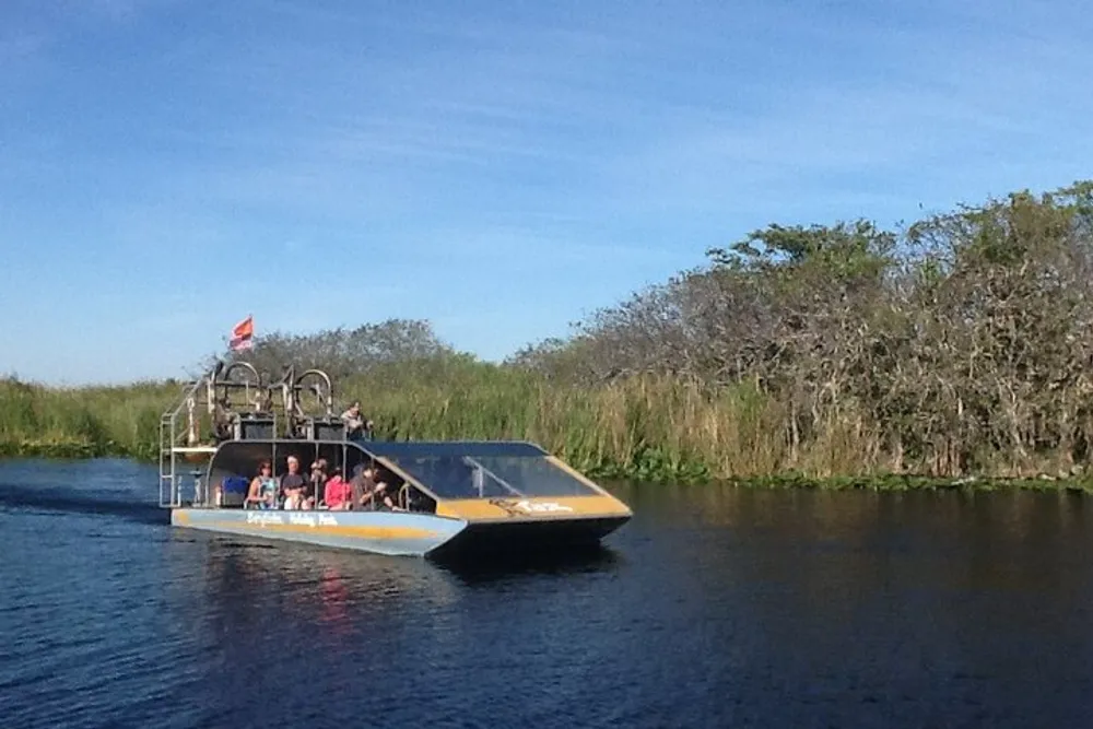 A group of tourists is enjoying a ride on an airboat through a calm waterway surrounded by dense vegetation under a clear sky