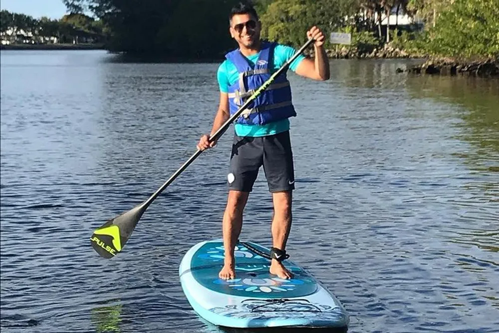 A man is smiling and standing on a stand-up paddleboard in calm water holding a paddle