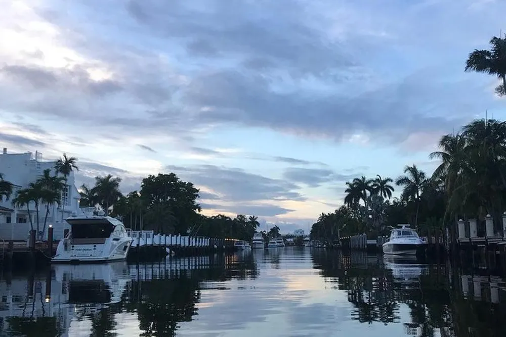 An evening view over a calm waterway flanked by private docks with moored boats and palm trees against a cloudy sky