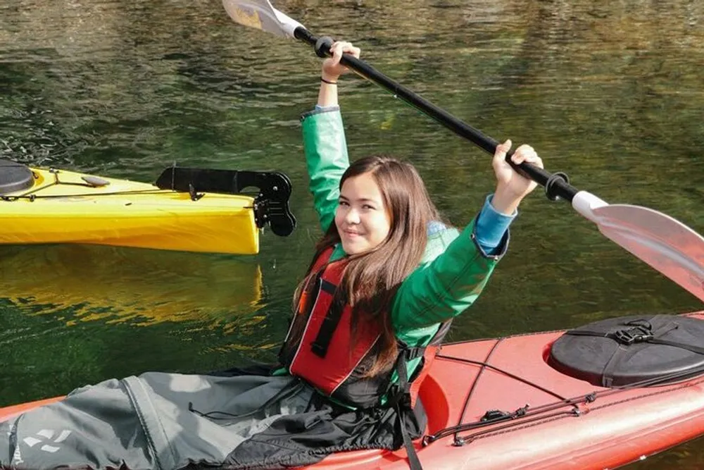 A smiling person is kayaking on calm water holding a paddle aloft