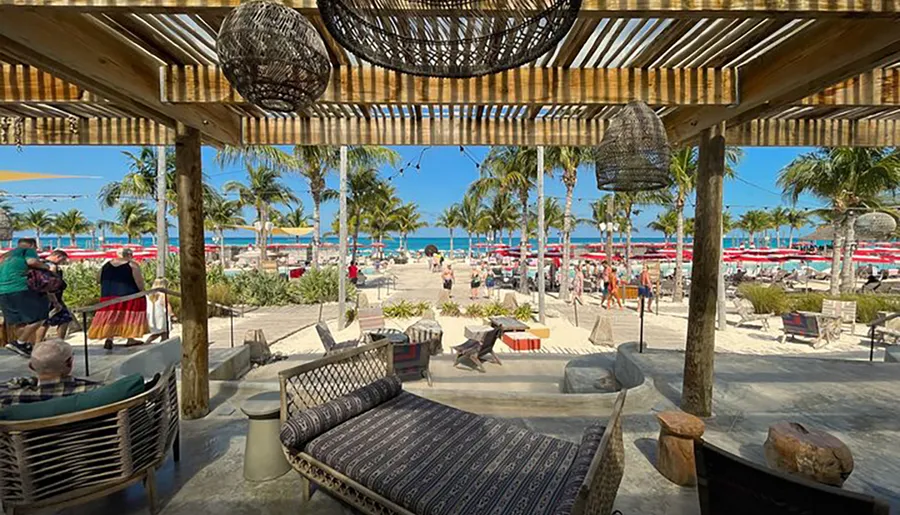 The image shows a tropical beachfront view from a shaded seating area with people enjoying the sunny day amidst palm trees and lounge chairs.