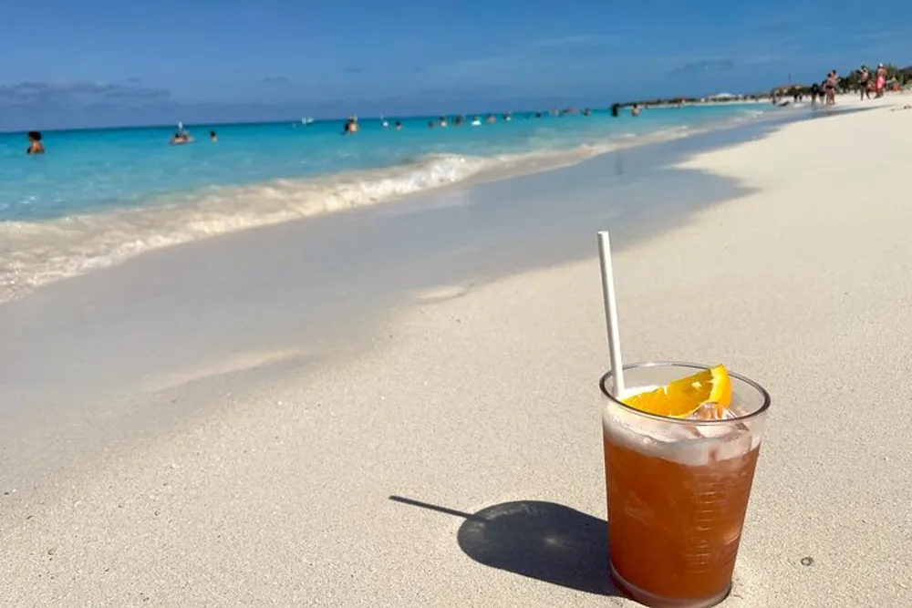 A refreshing drink with a slice of orange sits on a white sandy beach with the turquoise ocean and beachgoers in the background