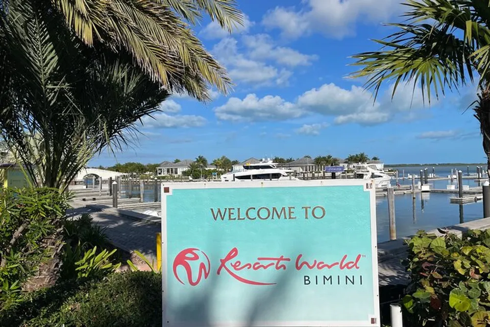 The image shows a Welcome to Resorts World Bimini sign with a view of palm trees a clear sky and a marina with boat slips in the background