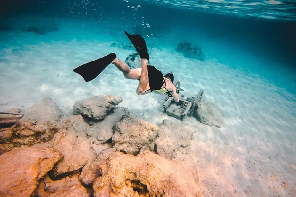 A diver with black fins is exploring the clear underwater environment near some rocky formations on the sandy sea bottom