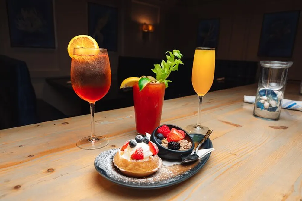 The image shows a beautifully arranged table with a variety of drinks including a garnished bloody mary a spritz a mimosa and a plate with a dessert topped with fresh berries and powdered sugar