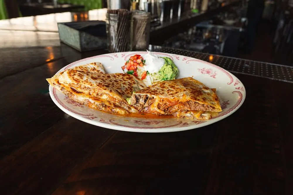 A golden brown quesadilla is sliced and served on a plate with a side of guacamole and sour cream on a wooden restaurant table