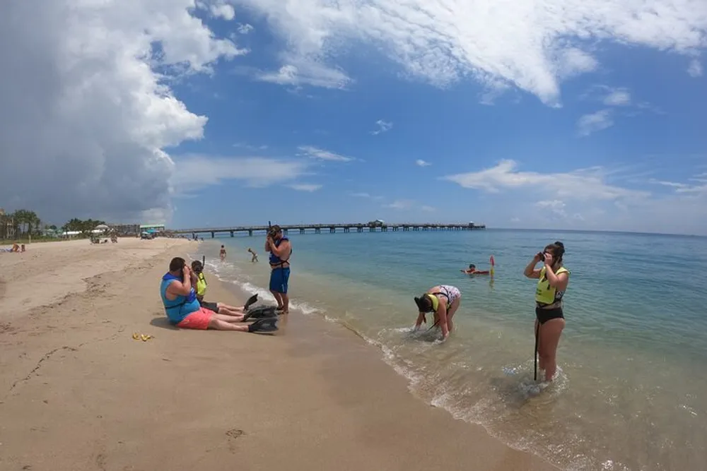 The image shows people enjoying a sunny day on a sandy beach with a pier extending into the calm sea in the background under a partially cloudy sky