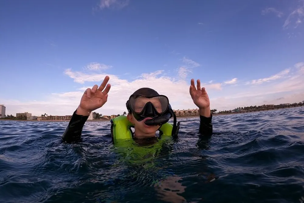 A person in a snorkeling mask and life vest is giving a peace sign with each hand while floating in the ocean