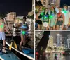 Two individuals are stand-up paddleboarding at night with LED lights on their boards creating a vibrant scene on the water against an urban backdrop