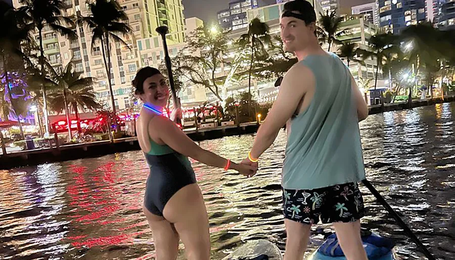 Two people are holding hands and standing on a paddleboard at night with an illuminated cityscape in the background.