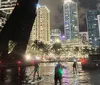 Two individuals are stand-up paddleboarding at night with LED lights on their boards creating a vibrant scene on the water against an urban backdrop