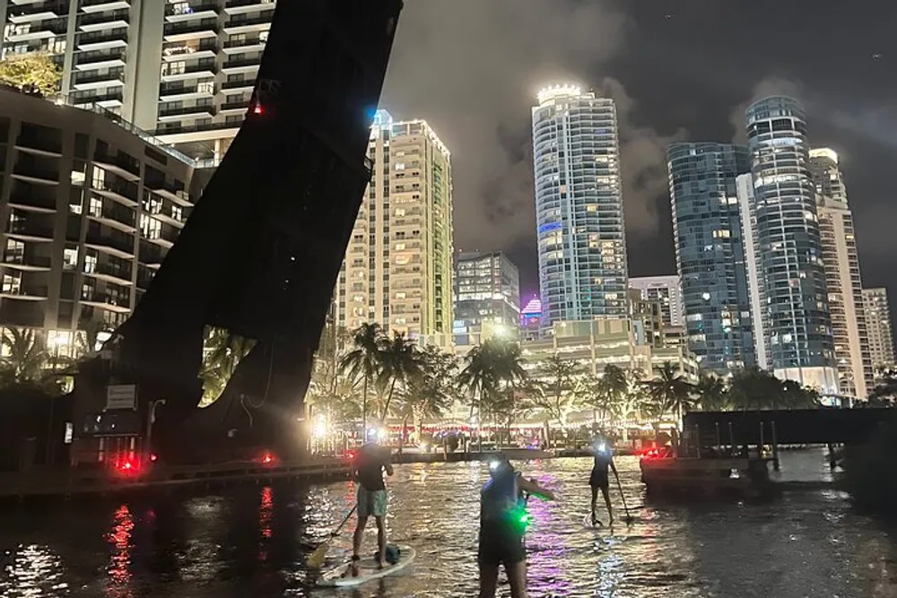 People are paddleboarding at night on a city river with illuminated skyscrapers in the background
