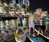 A man and a woman are holding hands while standing on paddleboards at night with city lights reflecting on the water in the background