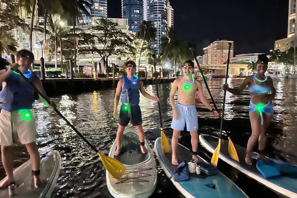Four individuals are standing on paddleboards at night equipped with glowing accessories in an urban waterfront setting