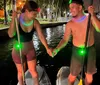 A man and a woman are holding hands while standing on paddleboards at night with city lights reflecting on the water in the background