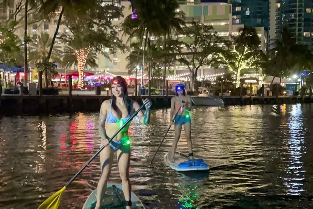 Two people are paddleboarding at night on an illuminated body of water with urban buildings in the background