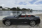 A black convertible car with red interior is parked by a waterfront with tropical vegetation in the background.