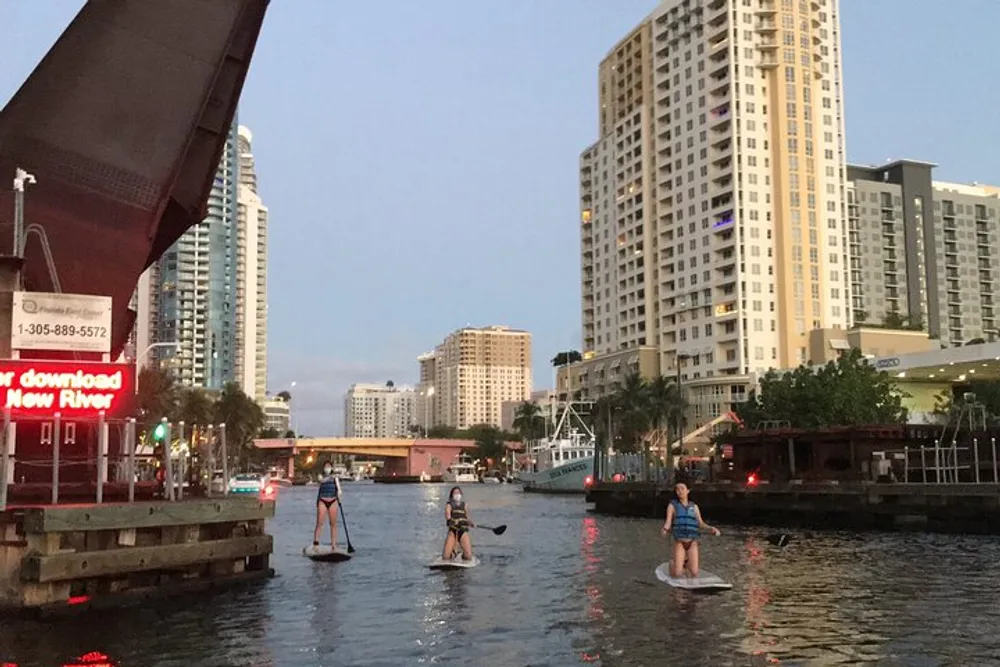 Three people are paddleboarding on an urban river against the backdrop of tall buildings during dusk