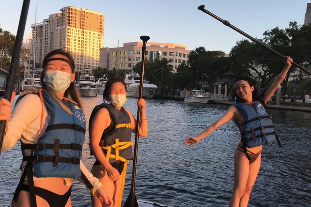 Three people wearing life jackets and face masks are holding paddles on a dock with a backdrop of a city waterfront bathed in golden sunlight