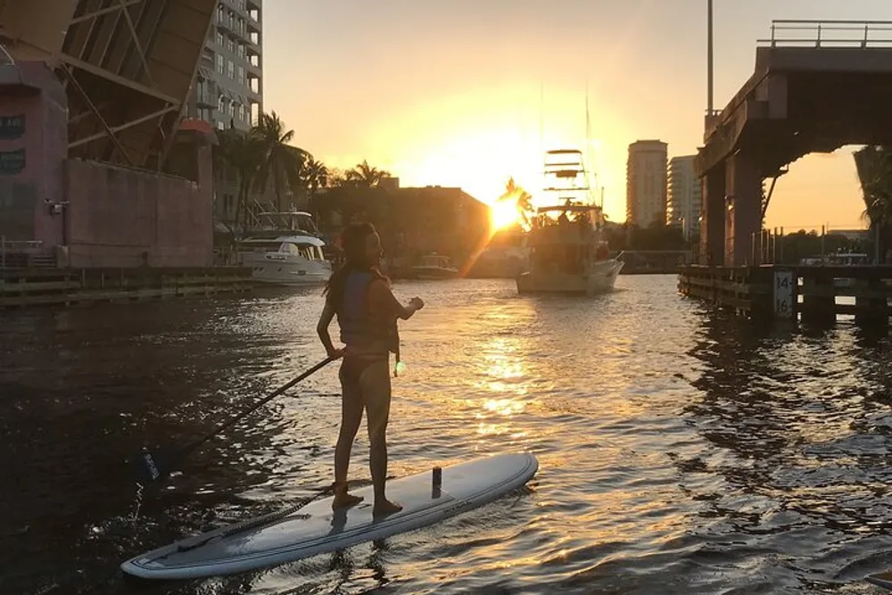 A person is stand-up paddleboarding on calm water during a beautiful sunset with a drawbridge and boats in the background